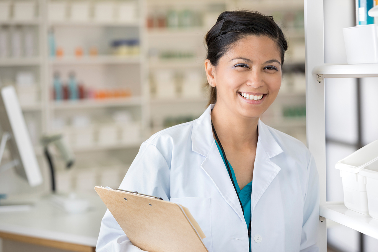 Smiling mid adult Asian female pharmacist stands in her pharmacy. She is smiling at the camera and is holding a clipboard.