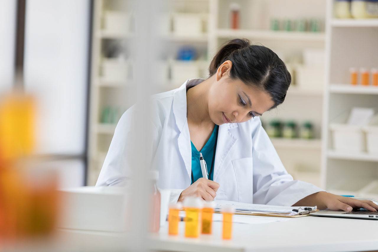 Mid adult female pharmacist concentrates while writing something on a clipboard. She is also using a laptop.